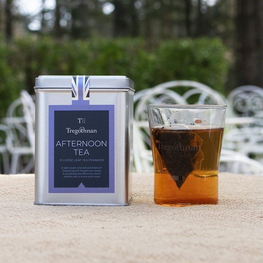 Tregothnan Afternoon Tea tin featuring loose leaf tea pyramids, with a glass tea cup filled with tea, set against a blurred garden background.